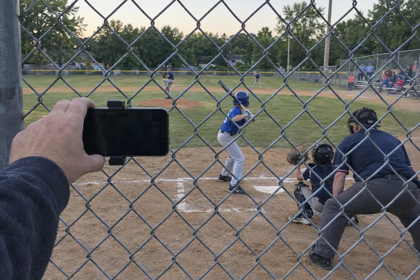 A dad clipping his phone to the backstop fence to stream a youth baseball game