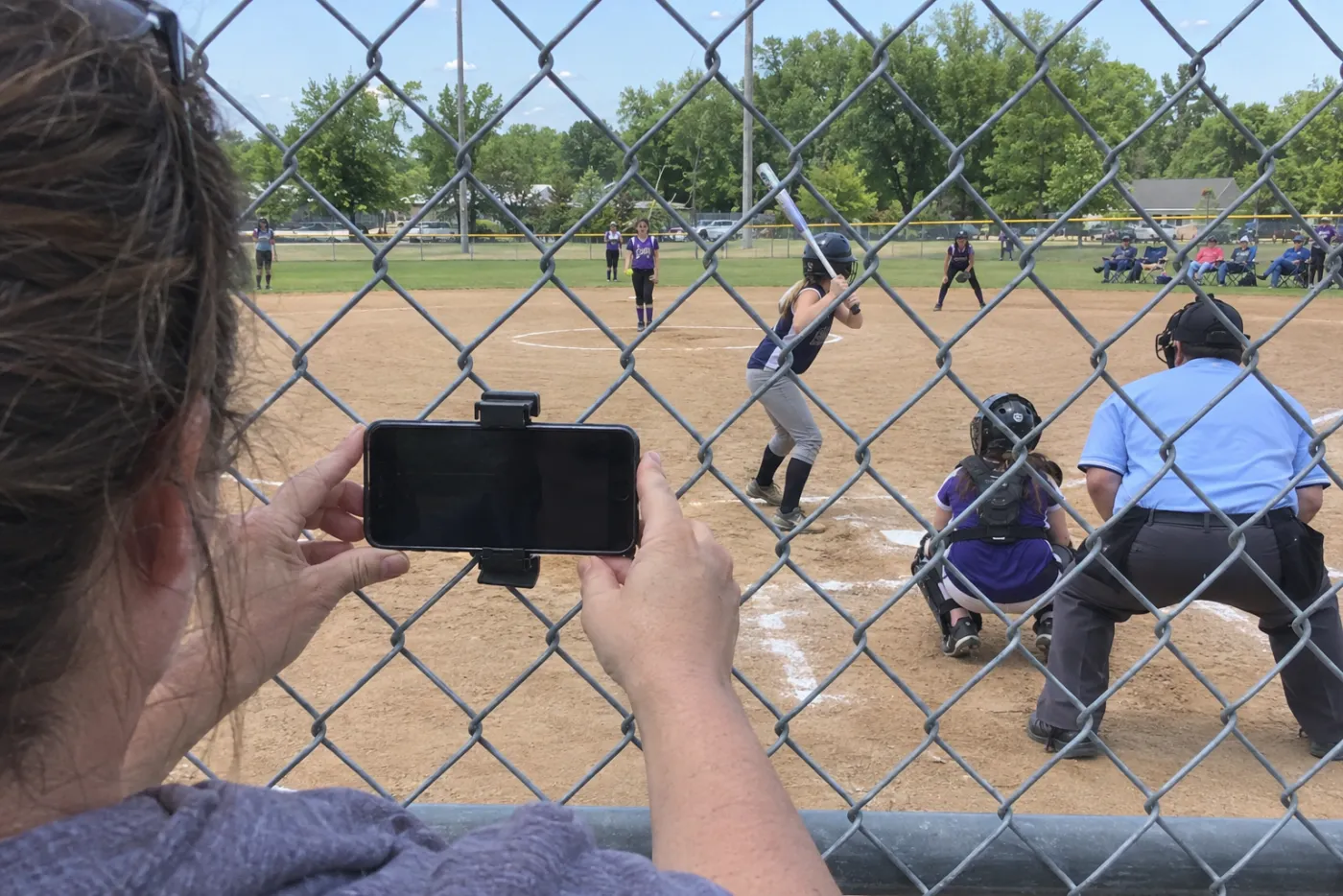 A parent clipping a phone to the backstop fence to stream a youth softball game